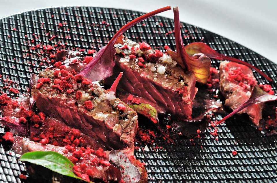 Beef course with red crumble and herb garnish on a black plate